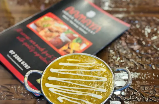 A small two-handled stainless steel dish of thick golden-green curry, drizzled with a zigzag of cream, standing on a dark carved wooden table. Behind it lies a red and black printed menu card showing photographs of fried snacks and a phone number.
