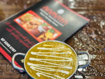 A small two-handled stainless steel dish of thick golden-green curry, drizzled with a zigzag of cream, standing on a dark carved wooden table. Behind it lies a red and black printed menu card showing photographs of fried snacks and a phone number.