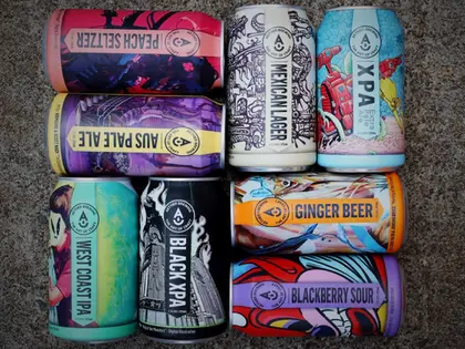 Eight beer cans laid flat in a grid on rough grey concrete, each wrapped in a different brightly coloured illustration. The labels read Peach Seltzer, Aus Pale Ale, West Coast IPA, Black XPA, Mexican Lager, XPA, Ginger Beer and Blackberry Sour.