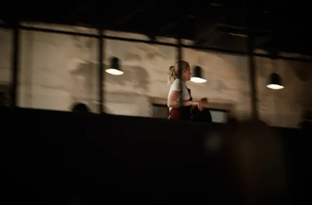 A dim, mostly shadowed interior seen over a dark parapet: a woman in a white T-shirt with a blonde ponytail sits side-on at a counter. Behind her a rough plaster wall, a row of small conical pendant lamps glowing warm, and a high steel-framed window.