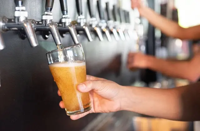 A hand holds a straight-sided glass beneath a chrome tap as pale golden beer pours in and a head of foam builds. A long row of taps runs away out of focus, and a second person fills another glass in the blurred background.