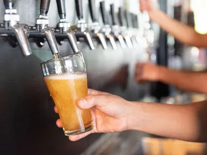 A hand holds a straight-sided glass beneath a chrome tap as pale golden beer pours in and a head of foam builds. A long row of taps runs away out of focus, and a second person fills another glass in the blurred background.