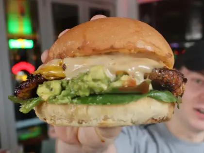 A person holding a burger up to the camera — a glossy brioche-style bun packed with charred grilled meat, smashed avocado, green leaves and a thick pale sauce — with red and pink neon signs glowing on the dark wall behind them.