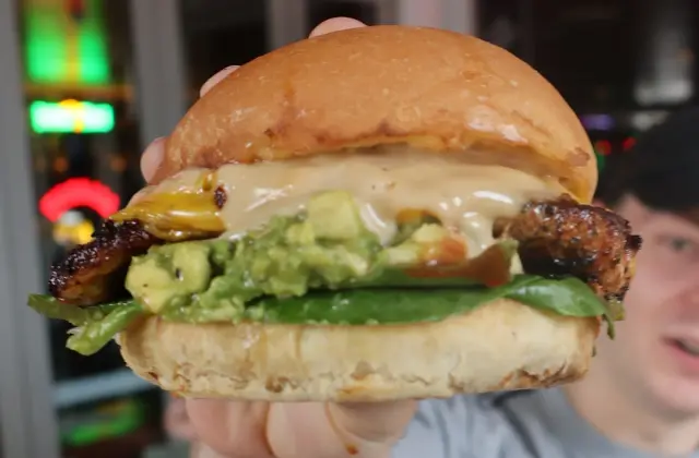 A person holding a burger up to the camera — a glossy brioche-style bun packed with charred grilled meat, smashed avocado, green leaves and a thick pale sauce — with red and pink neon signs glowing on the dark wall behind them.