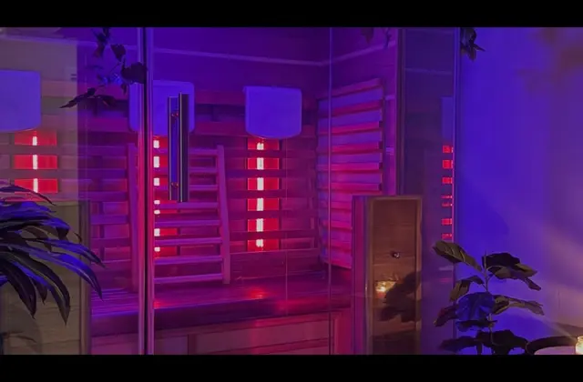 A timber-lined sauna cabin lit magenta and violet, its glass door open. Slatted pale wood walls carry glowing red heater panels and two rounded white fixtures, with a bench along the back. Leafy plants stand in the foreground and a candle burns on a small table at the right.