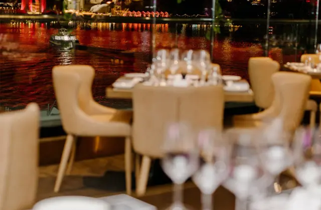 A dining room at night: pale upholstered armchairs around tables set with wine glasses and white napkins, behind a floor-to-ceiling window looking across the Brisbane River to the Story Bridge lit up in red, with a small boat passing on the water.