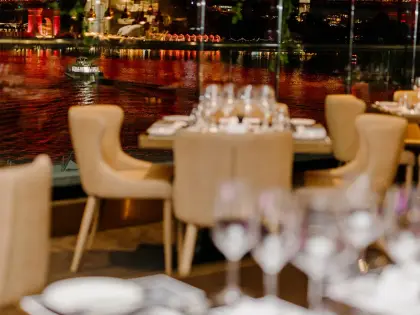 A dining room at night: pale upholstered armchairs around tables set with wine glasses and white napkins, behind a floor-to-ceiling window looking across the Brisbane River to the Story Bridge lit up in red, with a small boat passing on the water.