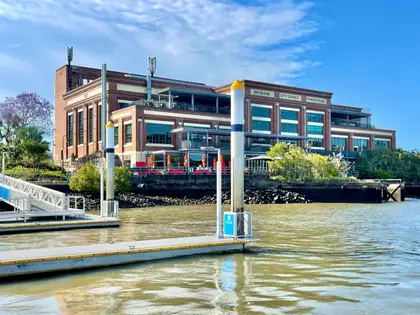 The red-brick former power station on the river bank, 'Brisbane City Council Power House' lettered across its upper wall, with glassed-in bars and red umbrellas along the terrace, seen across the water from a floating pontoon.