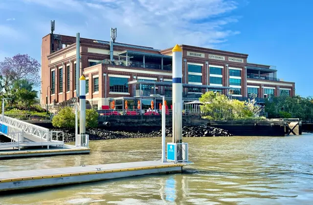 The red-brick former power station on the river bank, 'Brisbane City Council Power House' lettered across its upper wall, with glassed-in bars and red umbrellas along the terrace, seen across the water from a floating pontoon.