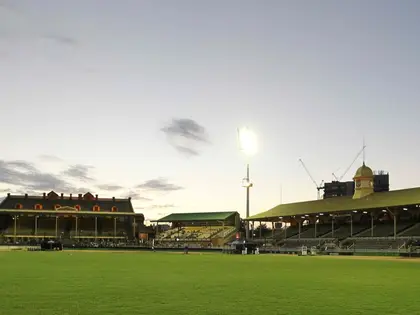 The showgrounds' main arena at dusk — a broad empty grass oval in front of two old grandstands, one brick and gabled, the other long and timber-roofed with a domed clock tower, a floodlight burning above them.