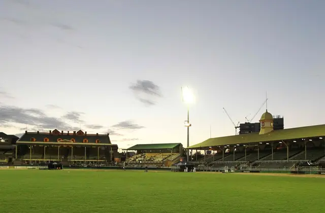 The showgrounds' main arena at dusk — a broad empty grass oval in front of two old grandstands, one brick and gabled, the other long and timber-roofed with a domed clock tower, a floodlight burning above them.