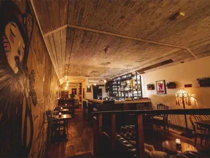 A long narrow room with a rough timber-boarded ceiling and dark floorboards, lit low and warm. A large hand-drawn mural of a woman's face with closed eyes covers the left wall; the right wall holds framed pictures, wall-mounted planters and a stained-glass lamp. Small tables with metal chairs line one side, leading to a bar backed by shelves of bottles, with a buttoned leather sofa in the foreground.