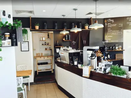 A small cafe counter: a chrome espresso machine, grinder and jug sit on a dark timber bench above white hexagonal tiling, with three enamel pendant lamps hanging above. The dark back wall carries a chalkboard menu headed "Clay", listing white coffees, black coffees, nitrogen and filter. Trailing pot plants, shelves of takeaway cups and a small white table and chairs fill the left of the frame.