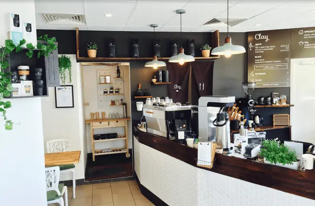 A small cafe counter: a chrome espresso machine, grinder and jug sit on a dark timber bench above white hexagonal tiling, with three enamel pendant lamps hanging above. The dark back wall carries a chalkboard menu headed "Clay", listing white coffees, black coffees, nitrogen and filter. Trailing pot plants, shelves of takeaway cups and a small white table and chairs fill the left of the frame.