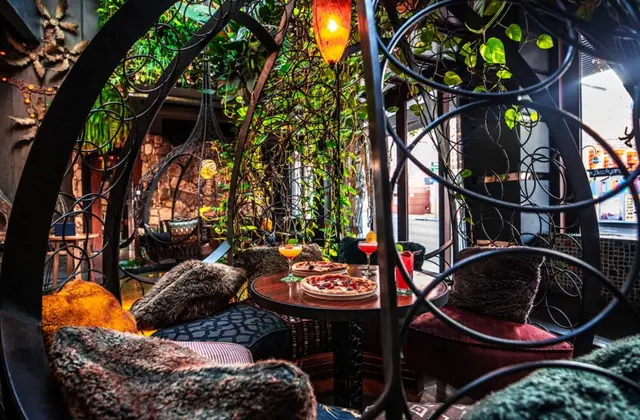 A seating nook framed by curling black wrought-iron scrollwork, with trailing vines overhead and a red glass pendant lamp. Two pizzas on white plates and three cocktails stand on a small round table between armchairs heaped with fur cushions; hanging egg chairs and a daylit street view sit beyond.