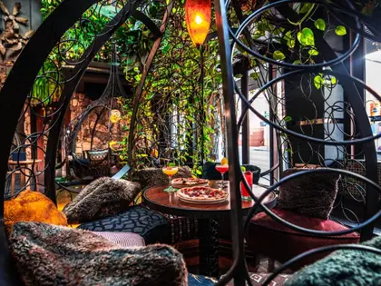 A seating nook framed by curling black wrought-iron scrollwork, with trailing vines overhead and a red glass pendant lamp. Two pizzas on white plates and three cocktails stand on a small round table between armchairs heaped with fur cushions; hanging egg chairs and a daylit street view sit beyond.