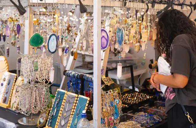 A jewellery stall hung with rows of polished agate and crystal pendants, with beaded necklaces on display boards and a shopper in a grey t-shirt browsing trays of rings.