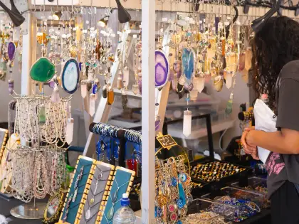 A jewellery stall hung with rows of polished agate and crystal pendants, with beaded necklaces on display boards and a shopper in a grey t-shirt browsing trays of rings.