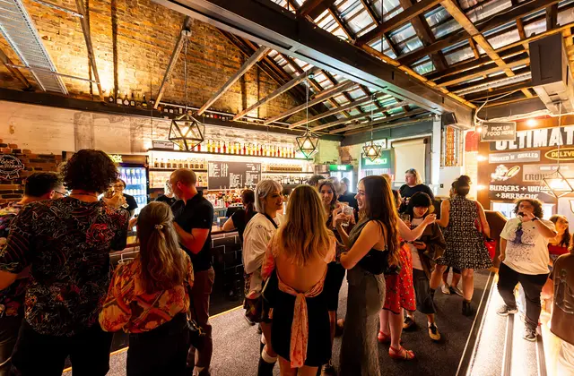 Patrons standing and talking with drinks in front of the bar, under a pitched roof of exposed timber beams and translucent sheeting with faceted pendant lamps; a chalkboard drinks list sits behind the bar and an illuminated food-counter sign is on the right.