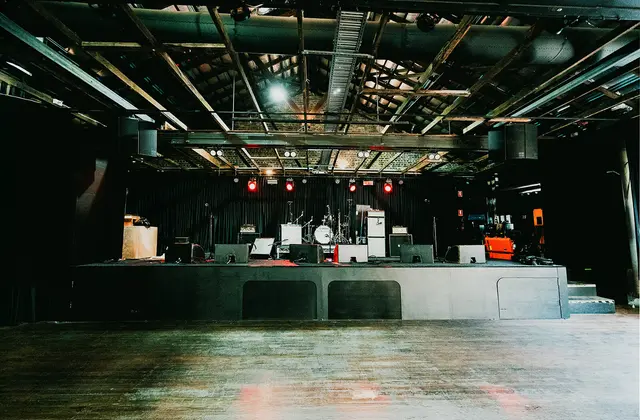 The empty band room seen from the floor: a low concrete stage set with a drum kit, guitar amps and wedge monitors in front of black curtains, red par lights above, and exposed timber roof trusses and a line-array speaker overhead.