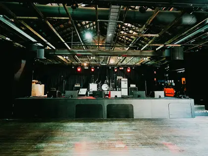 The empty band room seen from the floor: a low concrete stage set with a drum kit, guitar amps and wedge monitors in front of black curtains, red par lights above, and exposed timber roof trusses and a line-array speaker overhead.