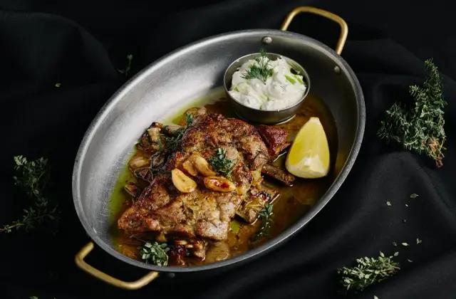 An oval metal pan with brass handles holding a browned, herb-crusted cut of meat on the bone in golden juices, with whole roasted garlic cloves, thyme sprigs, a lemon wedge and a small cup of thick white herbed yoghurt. The pan rests on rumpled black cloth scattered with loose thyme and oregano.