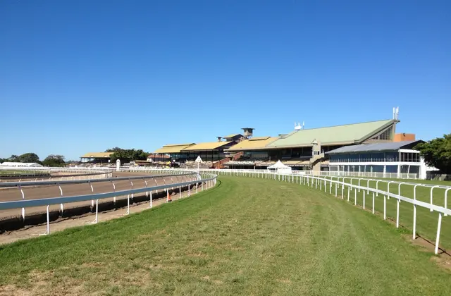 The empty turf track curving away toward the racecourse's long row of grandstands, with white running rails and the bare inner training track in the foreground under a cloudless sky.