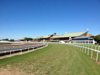 The empty turf track curving away toward the racecourse's long row of grandstands, with white running rails and the bare inner training track in the foreground under a cloudless sky.
