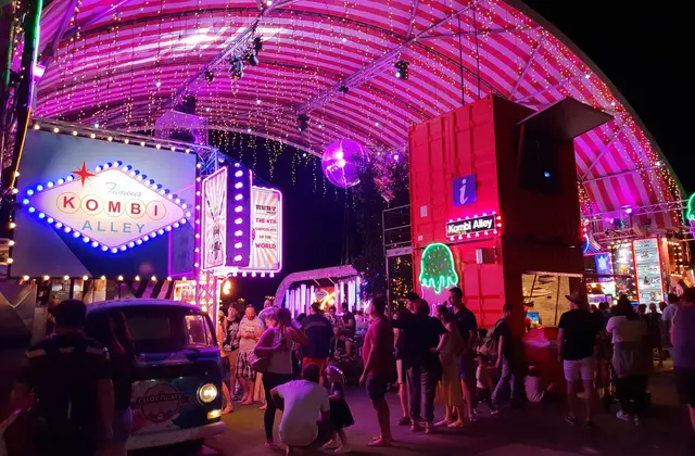 A night market walkway under a pink and white striped canopy strung with fairy lights and a mirror ball, a glowing 'Kombi Alley' sign on the left, a red shipping-container information booth on the right, and crowds around a Kombi van serving chocolate.