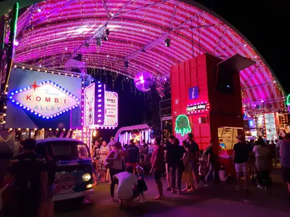 A night market walkway under a pink and white striped canopy strung with fairy lights and a mirror ball, a glowing 'Kombi Alley' sign on the left, a red shipping-container information booth on the right, and crowds around a Kombi van serving chocolate.