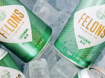 Four green and white beer cans lying in crushed ice, beaded with condensation. The labels read "Felons Brewing Co. Natural Ale", 355ml, 4.4% alcohol, above a small line-drawn emblem of hills and water inside a shield.