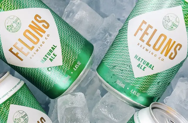 Four green and white beer cans lying in crushed ice, beaded with condensation. The labels read "Felons Brewing Co. Natural Ale", 355ml, 4.4% alcohol, above a small line-drawn emblem of hills and water inside a shield.
