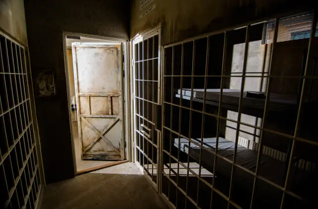 Inside a prison-cell escape room set: white-painted steel bars run floor to ceiling on both sides of a bare concrete corridor, a battered cross-braced timber door stands ajar spilling light in, and a stripped bunk bed with two thin mattresses sits behind the bars to the right.