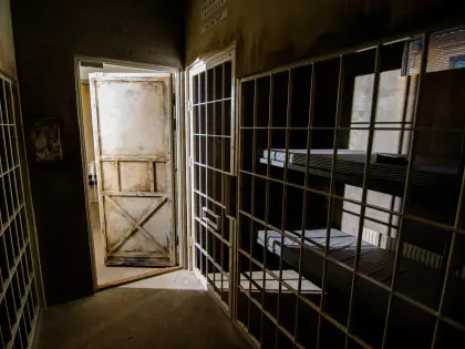 Inside a prison-cell escape room set: white-painted steel bars run floor to ceiling on both sides of a bare concrete corridor, a battered cross-braced timber door stands ajar spilling light in, and a stripped bunk bed with two thin mattresses sits behind the bars to the right.