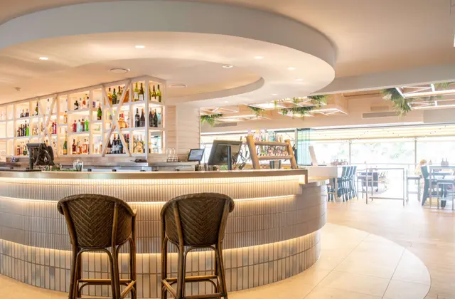 A curved bar in a bright white room, its front clad in fluted pale tile with a strip of light running along the base. Backlit shelving behind holds spirits and wine, and layered circular bulkheads ring the ceiling. Two rattan bar stools stand at the counter; beyond, a tiled floor gives onto timber tables and black chairs by open windows, with hanging greenery above.