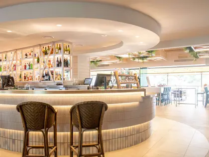 A curved bar in a bright white room, its front clad in fluted pale tile with a strip of light running along the base. Backlit shelving behind holds spirits and wine, and layered circular bulkheads ring the ceiling. Two rattan bar stools stand at the counter; beyond, a tiled floor gives onto timber tables and black chairs by open windows, with hanging greenery above.