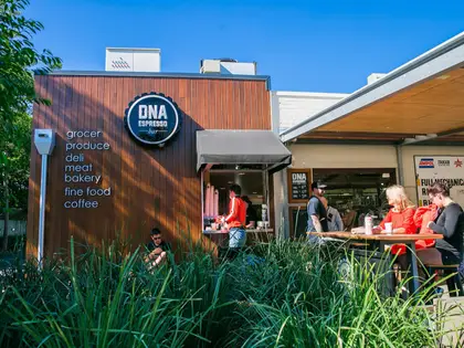 A single-storey building clad in dark vertical timber under a clear blue sky, carrying a round metal sign that reads "DNA Espresso Bar" and, beside it, a painted list: grocer, produce, deli, meat, bakery, fine food, coffee. A staff member works at an open serving window with a small awning; to the right, people sit at a timber table under a roofed area in front of a workshop hung with fuel-brand signs. Tall ornamental grasses fill the foreground and a man sits on a low ledge among them.