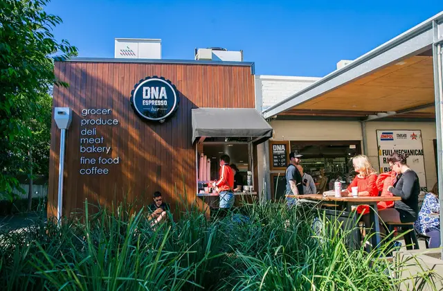 A single-storey building clad in dark vertical timber under a clear blue sky, carrying a round metal sign that reads "DNA Espresso Bar" and, beside it, a painted list: grocer, produce, deli, meat, bakery, fine food, coffee. A staff member works at an open serving window with a small awning; to the right, people sit at a timber table under a roofed area in front of a workshop hung with fuel-brand signs. Tall ornamental grasses fill the foreground and a man sits on a low ledge among them.