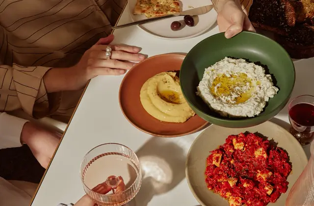 Looking down on a white table in hard sunlight. A woman in a tan pinstriped shirt holds a knife over a plate of flatbread and two olives, while a second pair of hands lowers a green bowl of thick white dip pooled with olive oil. Around them: a terracotta bowl of pale yellow puree, a plate of feta baked in red tomato sauce, a basket of dark charred toast, a glass of red wine and a ribbed glass holding a pale pink drink.
