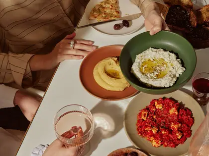 Looking down on a white table in hard sunlight. A woman in a tan pinstriped shirt holds a knife over a plate of flatbread and two olives, while a second pair of hands lowers a green bowl of thick white dip pooled with olive oil. Around them: a terracotta bowl of pale yellow puree, a plate of feta baked in red tomato sauce, a basket of dark charred toast, a glass of red wine and a ribbed glass holding a pale pink drink.