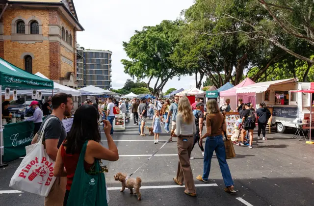 Crowds walking a line of white marquee stalls and a pink-awninged food van set up across a bitumen car park beside a two-storey brick building and large shade trees; a small poodle on a lead in the foreground.
