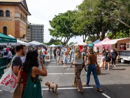 Crowds walking a line of white marquee stalls and a pink-awninged food van set up across a bitumen car park beside a two-storey brick building and large shade trees; a small poodle on a lead in the foreground.