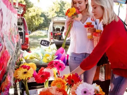 Two women at a flower stall, one smelling a bunch of orange and yellow gerberas while the other reaches into a tray of blooms; a chalkboard reads "$12" and a van is open behind them.