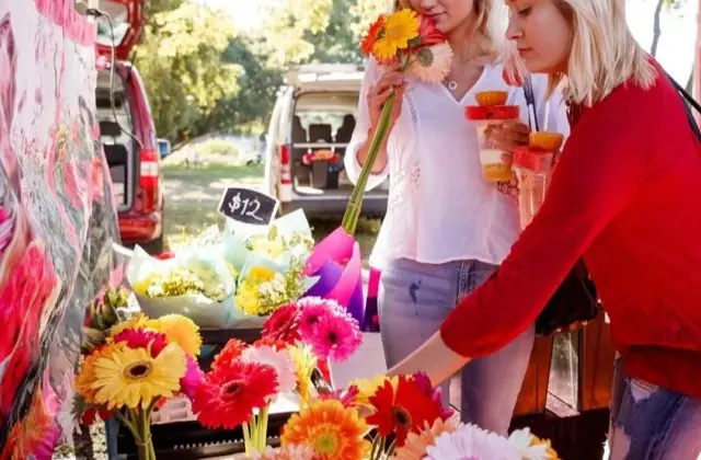 Two women at a flower stall, one smelling a bunch of orange and yellow gerberas while the other reaches into a tray of blooms; a chalkboard reads "$12" and a van is open behind them.