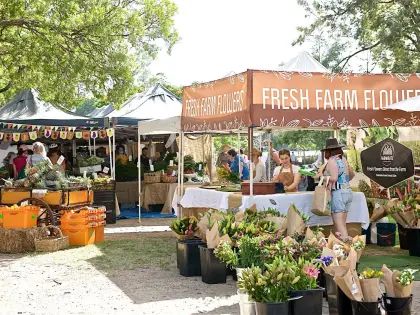 Buckets of wrapped flower bunches in the foreground of a market stall under an orange marquee printed "FRESH FARM FLOWERS", with shoppers browsing produce trestles under trees behind.