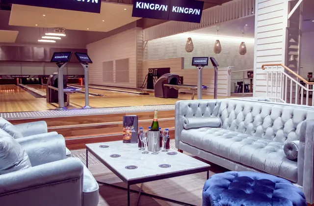 A lounge set inside a bowling alley: a buttoned pale-blue velvet sofa, armchair and ottoman around a marble coffee table holding a bottle of sparkling wine in a steel ice bucket and two flutes, with polished timber lanes, ball returns and screens on stands just beyond, under hanging signs reading KINGPIN.