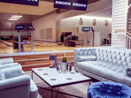A lounge set inside a bowling alley: a buttoned pale-blue velvet sofa, armchair and ottoman around a marble coffee table holding a bottle of sparkling wine in a steel ice bucket and two flutes, with polished timber lanes, ball returns and screens on stands just beyond, under hanging signs reading KINGPIN.