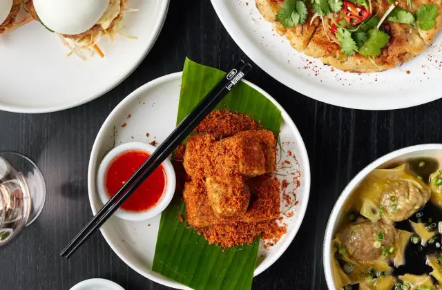 Overhead view of dishes crowded onto a black timber table: steamed bao buns filled with something crumbed and shredded slaw, a large golden fried pancake heaped with coriander, sliced red chilli and spring onion, chilli-crumbed tofu on a banana leaf with black chopsticks and a dish of sweet chilli sauce, a bowl of wontons and meatballs in dark broth, a herb salad topped with fried shallots, and a small dish of sliced green chilli.
