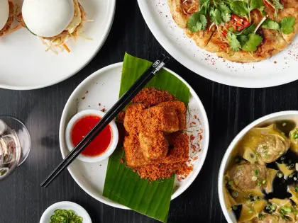 Overhead view of dishes crowded onto a black timber table: steamed bao buns filled with something crumbed and shredded slaw, a large golden fried pancake heaped with coriander, sliced red chilli and spring onion, chilli-crumbed tofu on a banana leaf with black chopsticks and a dish of sweet chilli sauce, a bowl of wontons and meatballs in dark broth, a herb salad topped with fried shallots, and a small dish of sliced green chilli.