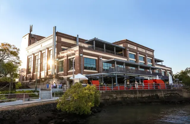The red-brick Brisbane Powerhouse seen from the river at golden hour, with its lettered parapet, steel chimneys, and a covered ground-floor terrace of diners under umbrellas along the riverside wall.
